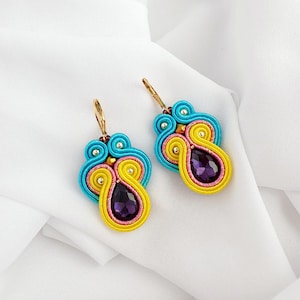 May include: A pair of dangle earrings with a yellow, pink and blue design. The earrings feature a purple teardrop-shaped gemstone in the centre.