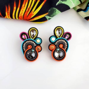Op de afbeelding: Een paar kleurrijke oorbellen met zwarte, oranje, blauwe, gele en roze accenten. De oorbellen hebben een groot, facetgeslepen, donkergrijs kristal in het midden van elke oorbel.