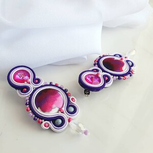 May include: A pair of purple, white, and pink earrings with crystal accents. The earrings are made with a technique called peyote stitch, which creates a textured, three-dimensional effect.