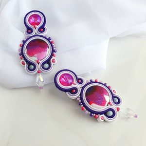 May include: A pair of purple and white earrings with a pink and white patterned center. The earrings are made with small beads and have a crystal dangle.