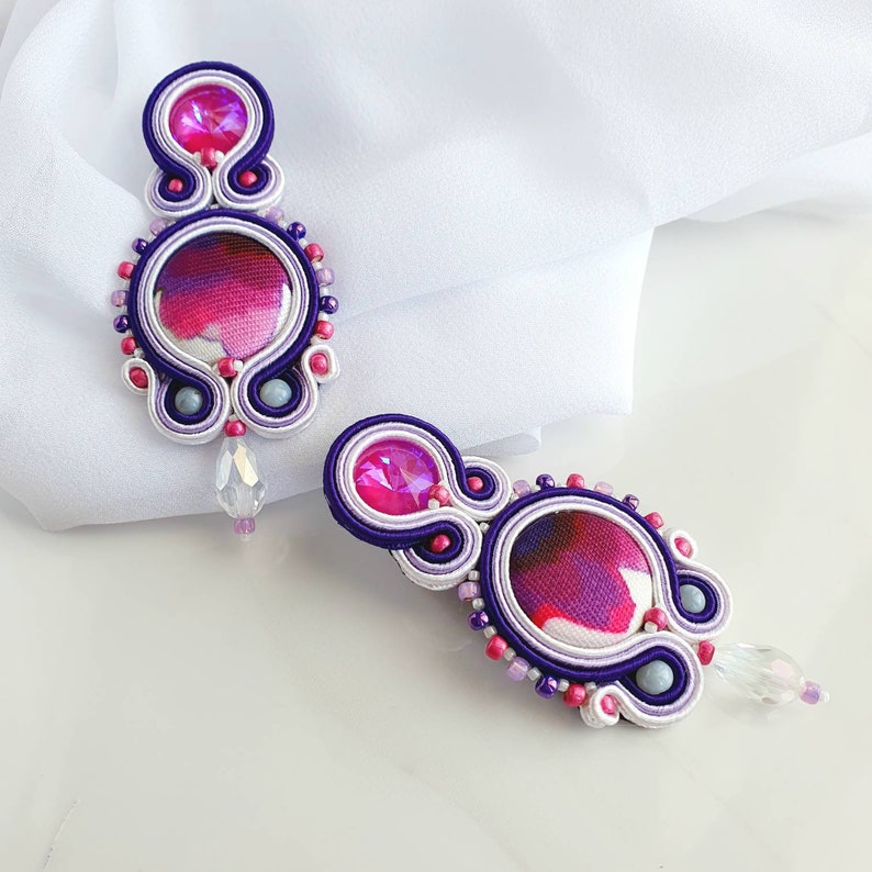 May include: A pair of purple and white earrings with a pink and white patterned center. The earrings are made with small beads and have a crystal dangle.