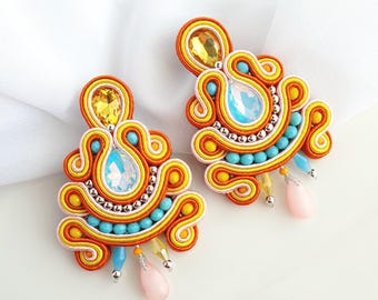 May include: A pair of orange, yellow, and turquoise earrings with a teardrop-shaped crystal in the center. The earrings have a beaded fringe and a pink stone at the bottom.