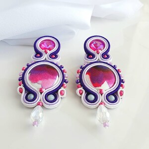 May include: A pair of white, purple, and pink earrings with a round, colorful fabric center. The earrings have a crystal-like bead hanging from the bottom.