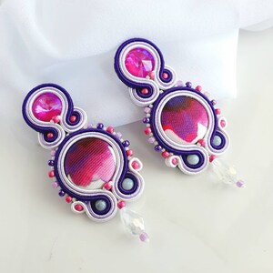 May include: A pair of dangle earrings with a purple, pink, and white swirl design. The earrings feature a round, patterned fabric center and a crystal accent.