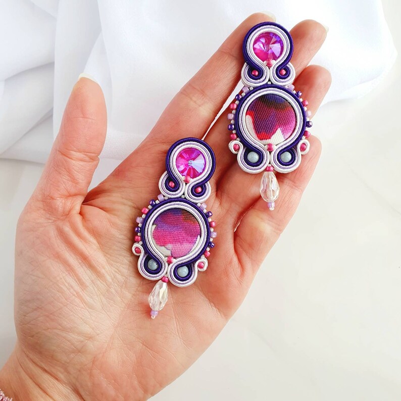 May include: A pair of purple and white earrings with pink and white fabric accents and clear crystal drops.