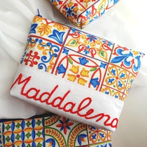 May include: A canvas pouch with a vibrant tile design in blue, yellow, and red. The name "Maddalena" is embroidered in red across the bottom. Another pouch with a similar pattern is visible.
