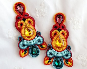 May include: A pair of colorful earrings with red, orange, yellow, green, and blue thread wrapped around a central yellow and clear rhinestone. The earrings have a teardrop shape and are accented with smaller rhinestones.