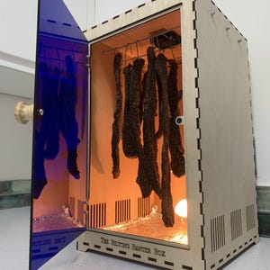 Op de afbeelding: Een houten doos met een blauwe deur en een lampje erin. De doos is gelabeld "The Belting Banger Box". Er hangen reepjes gedroogd vlees in de doos.