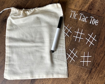 May include: A white drawstring bag, a black dry-erase marker, and a clear acrylic Tic Tac Toe game board with white lines. The board has the words "Tic Tac Toe" printed on it.