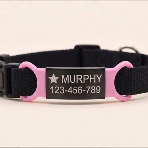 May include: A black nylon pet collar featuring a central rectangular black metal identification tag with a star icon, the name MURPHY, and the phone number 123-456-789. The tag is held by two pink silicone loops and includes a black plastic buckle.