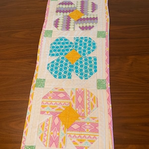 May include: A white table runner with three colorful flower designs. The flowers are made from fabric with different patterns, including polka dots, geometric shapes, and stripes. The flowers are in shades of purple, blue, pink, yellow, and green.