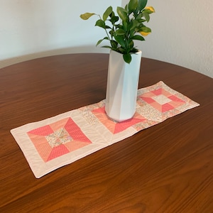 May include: A pink and white quilted table runner with a floral pattern. The runner is on a wooden table with a white vase and green plant.