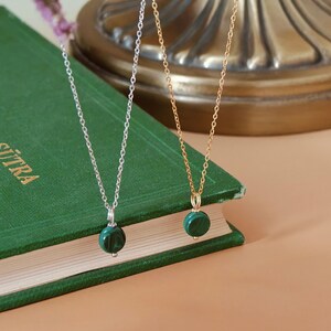 Peut inclure: Deux colliers avec des pendentifs ronds en malachite verte. L'un a une cha&icirc;ne argent&eacute;e, l'autre une cha&icirc;ne dor&eacute;e. Les pendentifs sont suspendus &agrave; de fines cha&icirc;nes. Les colliers reposent sur un livre vert.