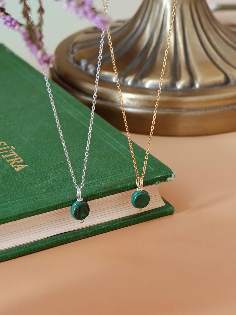 Peut inclure: Deux colliers avec des pendentifs ronds en malachite verte. L'un a une cha&icirc;ne argent&eacute;e, l'autre une cha&icirc;ne dor&eacute;e. Les pendentifs sont suspendus &agrave; de fines cha&icirc;nes. Les colliers reposent sur un livre vert.