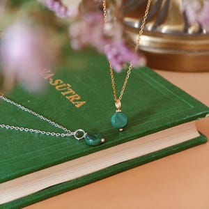 Peut inclure: Deux colliers avec des pendentifs en malachite reposent sur un livre vert. L'un a une cha&icirc;ne dor&eacute;e, l'autre une cha&icirc;ne argent&eacute;e. Le livre porte le texte "VA-SUTRA". L'arri&egrave;re-plan est flou avec des tons roses et dor&eacute;s.