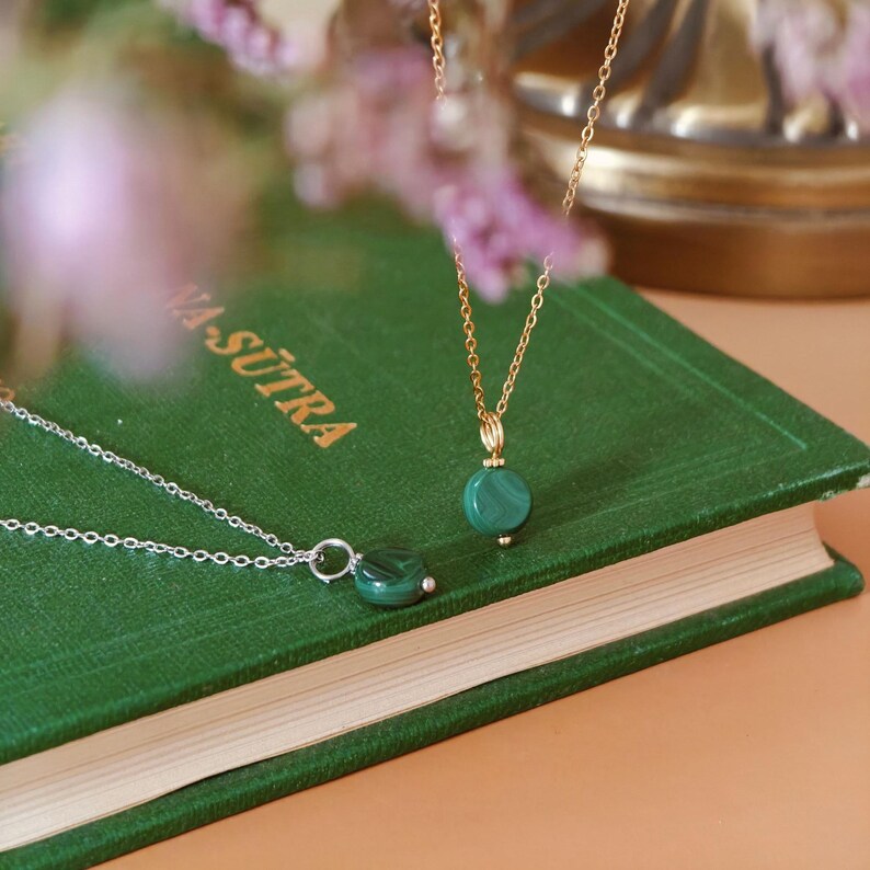 Peut inclure: Deux colliers avec des pendentifs en malachite reposent sur un livre vert. L'un a une cha&icirc;ne dor&eacute;e, l'autre une cha&icirc;ne argent&eacute;e. Le livre porte le texte "VA-SUTRA". L'arri&egrave;re-plan est flou avec des tons roses et dor&eacute;s.