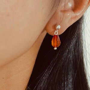 K&ouml;nnte beinhalten: Nahaufnahme eines silbernen Ohrrings mit einem tropfenf&ouml;rmigen, orange-roten Edelstein. Der Ohrring ist an einer kleinen Silberkugel am Ohrl&auml;ppchen befestigt. Der Edelstein hat eine facettierte Oberfl&auml;che.