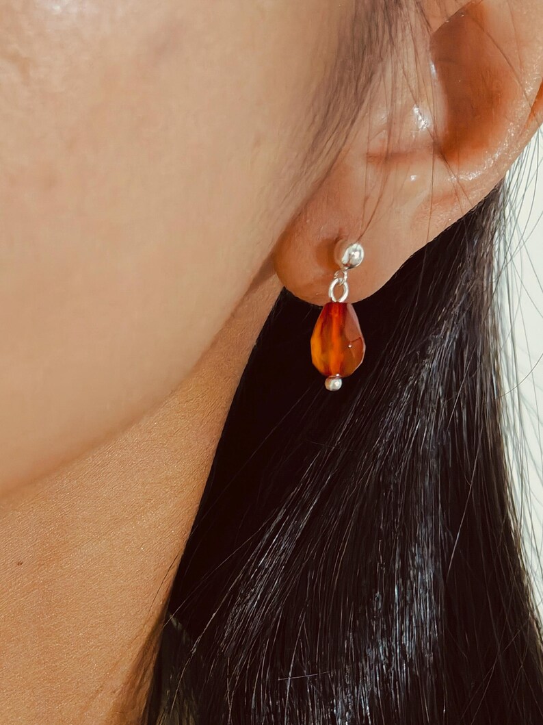 K&ouml;nnte beinhalten: Nahaufnahme eines silbernen Ohrrings mit einem tropfenf&ouml;rmigen, orange-roten Edelstein. Der Ohrring ist an einer kleinen Silberkugel am Ohrl&auml;ppchen befestigt. Der Edelstein hat eine facettierte Oberfl&auml;che.