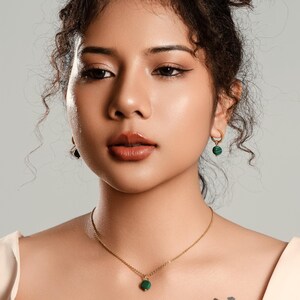 Peut inclure: Une femme porte un collier en or avec un petit pendentif rond vert. Elle porte &eacute;galement des boucles d'oreilles cr&eacute;oles dor&eacute;es avec des accents verts. La femme a les cheveux fonc&eacute;s et un tatouage de montagne et de fleurs sur la poitrine.