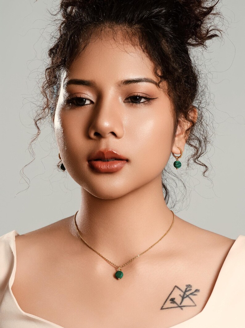 Peut inclure: Une femme porte un collier en or avec un petit pendentif rond vert. Elle porte &eacute;galement des boucles d'oreilles cr&eacute;oles dor&eacute;es avec des accents verts. La femme a les cheveux fonc&eacute;s et un tatouage de montagne et de fleurs sur la poitrine.