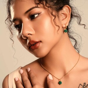 Peut inclure: Un collier en or et des boucles d'oreilles assorties avec des pierres rondes de malachite verte. Le collier a un petit pendentif. Les boucles d'oreilles sont de petits cerceaux avec la m&ecirc;me pierre verte. Les bijoux sont port&eacute;s par une personne.