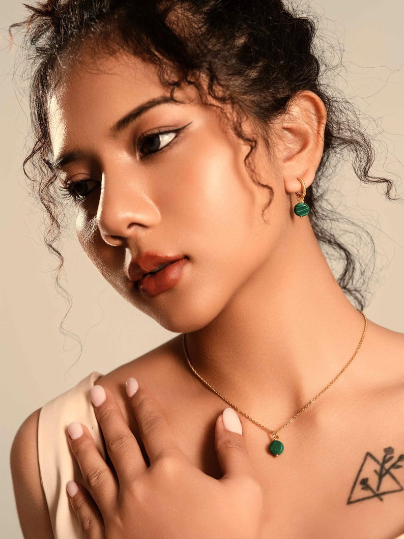 Peut inclure: Un collier en or et des boucles d'oreilles assorties avec des pierres rondes de malachite verte. Le collier a un petit pendentif. Les boucles d'oreilles sont de petits cerceaux avec la m&ecirc;me pierre verte. Les bijoux sont port&eacute;s par une personne.