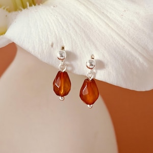 Può includere: Un paio di orecchini pendenti con perline ambrate sfaccettate a forma di goccia, sospese da perni color argento. Ogni orecchino ha una piccola perla d'argento nella parte inferiore della perla ambrata. Sfondo bianco.