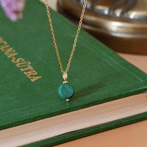Peut inclure: Un collier en cha&icirc;ne dor&eacute;e avec un pendentif rond en malachite verte. Le collier est pos&eacute; sur un livre vert avec des lettres dor&eacute;es. Le pendentif est vert fonc&eacute; avec une b&eacute;li&egrave;re dor&eacute;e.