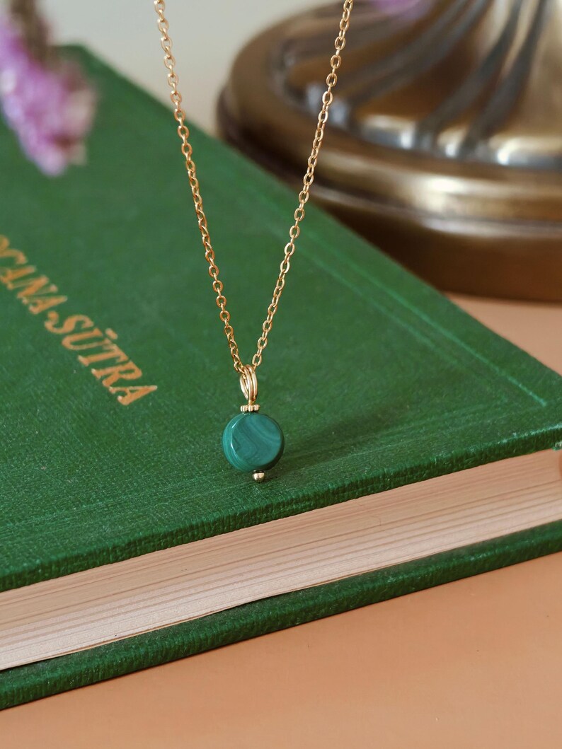 Peut inclure: Un collier en cha&icirc;ne dor&eacute;e avec un pendentif rond en malachite verte. Le collier est pos&eacute; sur un livre vert avec des lettres dor&eacute;es. Le pendentif est vert fonc&eacute; avec une b&eacute;li&egrave;re dor&eacute;e.
