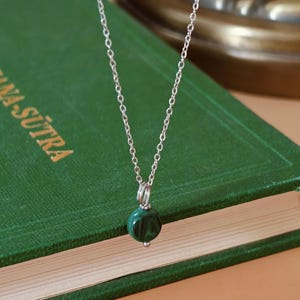 Peut inclure: Un collier en argent avec un petit pendentif rond en malachite vert fonc&eacute;. Le pendentif est suspendu &agrave; une cha&icirc;ne d&eacute;licate. Le collier est pr&eacute;sent&eacute; sur un livre vert.