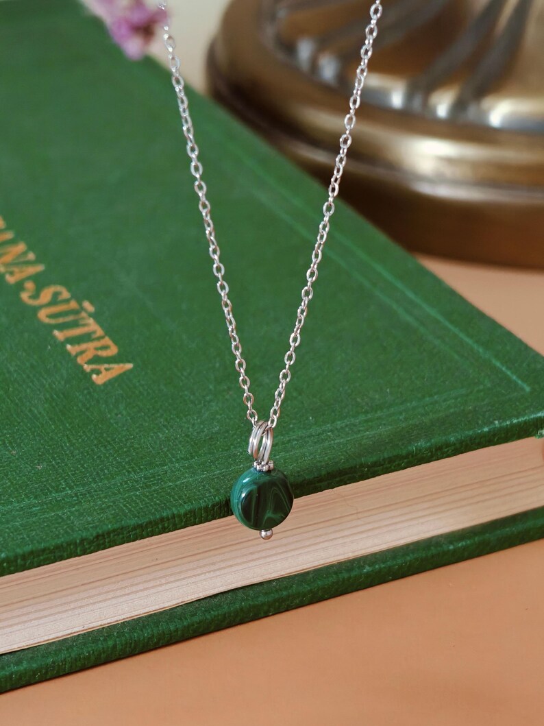 Peut inclure: Un collier en argent avec un petit pendentif rond en malachite vert fonc&eacute;. Le pendentif est suspendu &agrave; une cha&icirc;ne d&eacute;licate. Le collier est pr&eacute;sent&eacute; sur un livre vert.