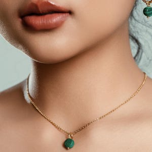 Peut inclure: Un collier dor&eacute; avec un petit pendentif rond vert. Le collier est d&eacute;licat et suspendu &agrave; une cha&icirc;ne. Une boucle d'oreille assortie avec une pierre verte est visible. Les bijoux sont sur un fond neutre.