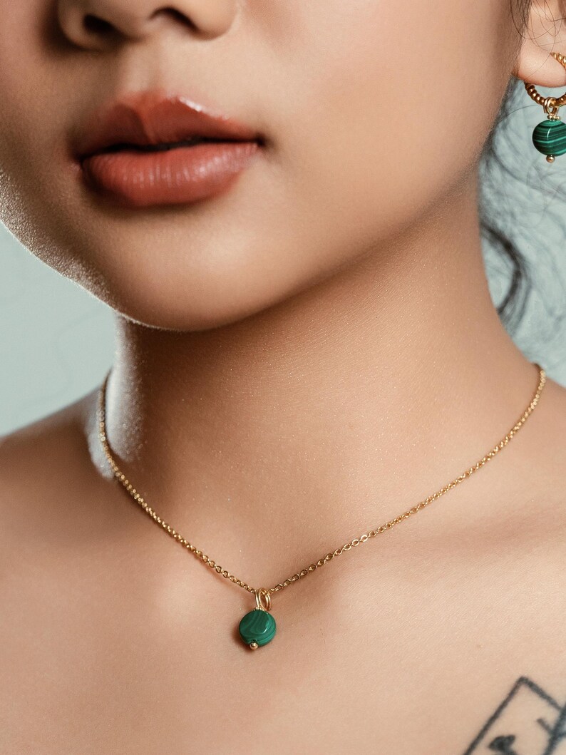 Peut inclure: Un collier dor&eacute; avec un petit pendentif rond vert. Le collier est d&eacute;licat et suspendu &agrave; une cha&icirc;ne. Une boucle d'oreille assortie avec une pierre verte est visible. Les bijoux sont sur un fond neutre.