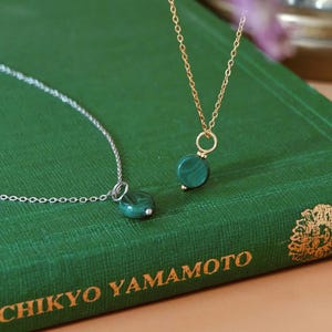 Peut inclure: Deux colliers avec des pendentifs en malachite verte. L'un a une cha&icirc;ne dor&eacute;e, l'autre une cha&icirc;ne argent&eacute;e. Les pendentifs ronds ont un aspect marbr&eacute;. Les colliers sont pr&eacute;sent&eacute;s sur un livre vert.