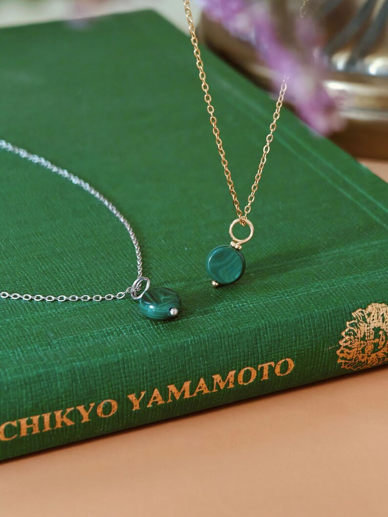 Peut inclure: Deux colliers avec des pendentifs en malachite verte. L'un a une cha&icirc;ne dor&eacute;e, l'autre une cha&icirc;ne argent&eacute;e. Les pendentifs ronds ont un aspect marbr&eacute;. Les colliers sont pr&eacute;sent&eacute;s sur un livre vert.