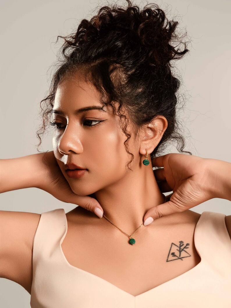 Peut inclure: Un collier en or et une paire de boucles d'oreilles assorties avec de petits pendentifs ronds en pierre verte. Le bijou est port&eacute; par une personne aux cheveux fonc&eacute;s. L'ensemble est assorti.