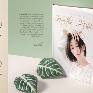 Pu&ograve; includere: Un libro bianco con il titolo "The Life Story of Ananda Khristie" in un font corsivo. Il libro &egrave; aperto per rivelare una foto di una donna con capelli neri corti. Il libro &egrave; appoggiato su una superficie bianca con due foglie verdi.