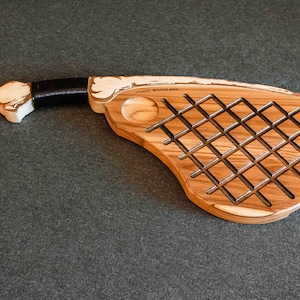 May include: A wooden serving board shaped like a racquet with a grid pattern and a small round indentation. The board is made of light brown wood with a white painted handle and black accents.