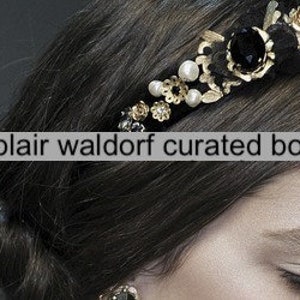 May include: A gold headband with black and white embellishments, including pearls, flowers, and a large black gem. The headband is worn in a woman's hair.