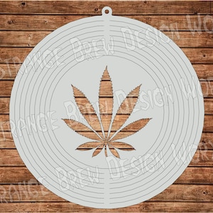 May include: A white circular metal wind spinner with a stylized cannabis leaf design. The spinner has concentric circles that create a swirling effect when it spins.