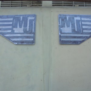 Puede incluir: Dos rejillas metálicas con la letra "M" recortada en el centro. Las rejillas están fijadas a una superficie verde.