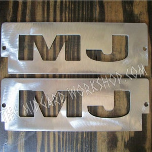 May include: Two silver metal plates with the letters "MJ" cut out. The plates are on a wooden surface. The text "THE MIDGARD WORKSHOP.COM" is visible on the bottom plate.