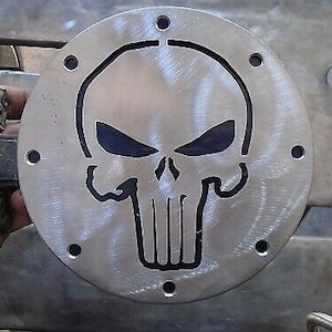 May include: A round metal skull design with a black outline. The skull has a menacing expression with a furrowed brow and a pointed chin. The design is cut out of a metal plate and has a brushed finish.