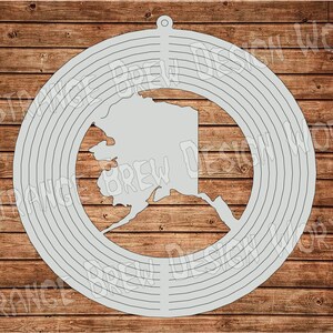 Peut inclure: Une silhouette grise de l'état d'Alaska à l'intérieur d'un motif circulaire avec des cercles concentriques. Le motif est sur un fond en bois.