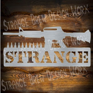 Puede incluir: Un letrero de metal con la palabra "STRANGE" en letras grandes y en negrita. Sobre la palabra hay una silueta de un rifle con una fila de balas debajo. El letrero tiene un aspecto rústico y desgastado.