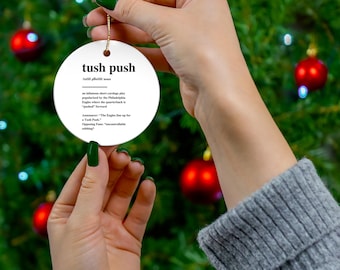 Op de afbeelding: Een witte, ronde kerstversiering met de zwarte tekst "tush push". De ornament heeft een gouden touwtje om op te hangen en wordt vastgehouden voor een kerstboom met rode ballen.