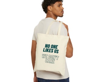 May include: A person is carrying a beige tote bag with the text "NO ONE LIKES US WE DON'T CARE" printed in dark teal.