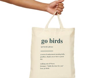 May include: A cream-colored canvas tote bag featuring dark green text that reads go birds, followed by a phonetic pronunciation, a definition as a term of endearment, and a short dialogue example involving Wawa and a stranger holding a door.