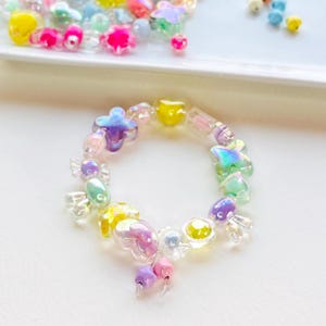 May include: A colorful bracelet made of translucent beads in various shapes, including flowers, hearts, and butterflies. The bracelet features a mix of pastel colors like yellow, pink, purple, and blue. Additional beads are scattered on a white surface.