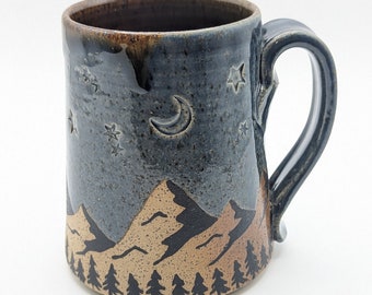 May include: A ceramic mug with a dark blue glaze, featuring a mountain and forest design. The mug has a crescent moon and star details. The base of the mug has a brown color, and the handle is curved.